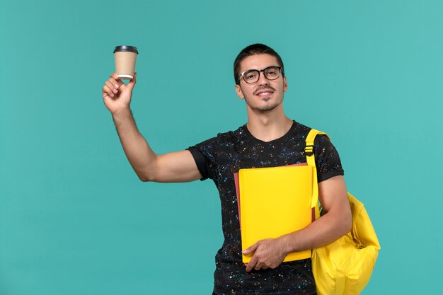 Vue de face de l'étudiant masculin en t-shirt noir sac à dos jaune tenant différents fichiers et café sur le mur bleu