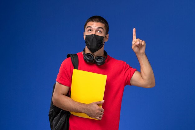 Vue de face étudiant masculin en t-shirt rouge portant un masque avec sac à dos tenant un fichier jaune sur le fond bleu.