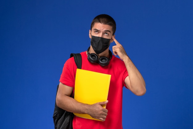 Vue de face étudiant masculin en t-shirt rouge portant un masque avec sac à dos tenant un fichier jaune pensant sur fond bleu.