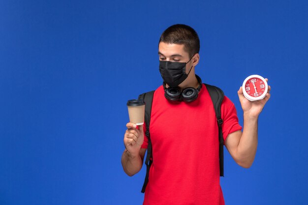 Vue de face étudiant masculin en t-shirt rouge portant un masque avec sac à dos tenant des horloges et du café sur fond bleu.