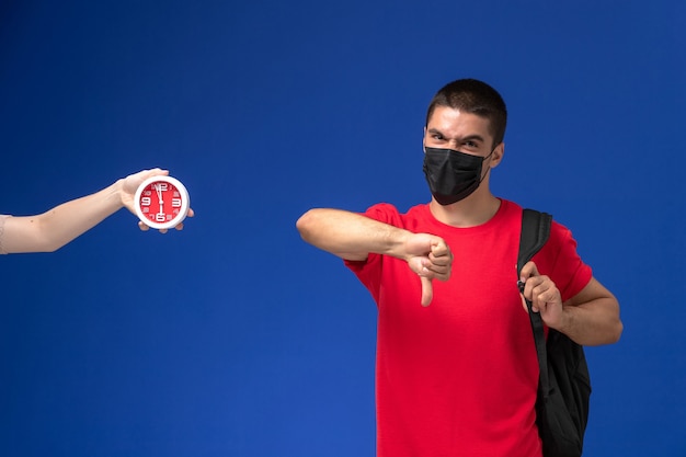 Vue de face étudiant masculin en t-shirt rouge portant un sac à dos avec masque posant sur le fond bleu.