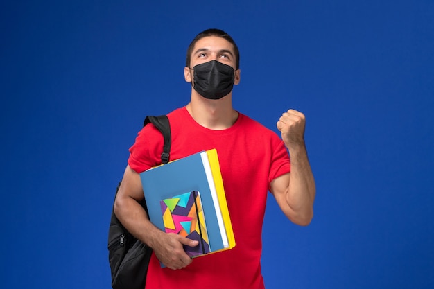 Vue de face étudiant masculin en t-shirt rouge portant un sac à dos en masque stérile noir tenant un cahier et des fichiers sur un bureau bleu.