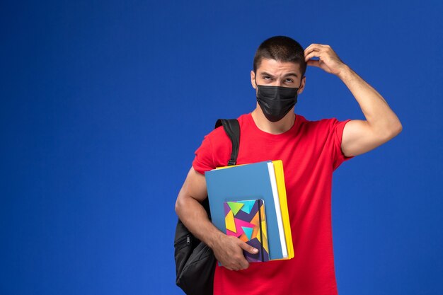 Vue de face étudiant masculin en t-shirt rouge portant sac à dos en masque stérile noir tenant des fichiers et de la pensée sur fond bleu.