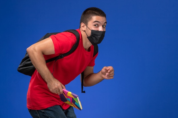 Vue de face étudiant masculin en t-shirt rouge portant un sac à dos en masque stérile noir tenant un stylo et un cahier en cours d'exécution sur un bureau bleu.