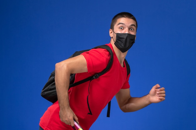 Vue de face étudiant masculin en t-shirt rouge portant un sac à dos en masque stérile noir tenant un stylo et un cahier en cours d'exécution sur fond bleu.