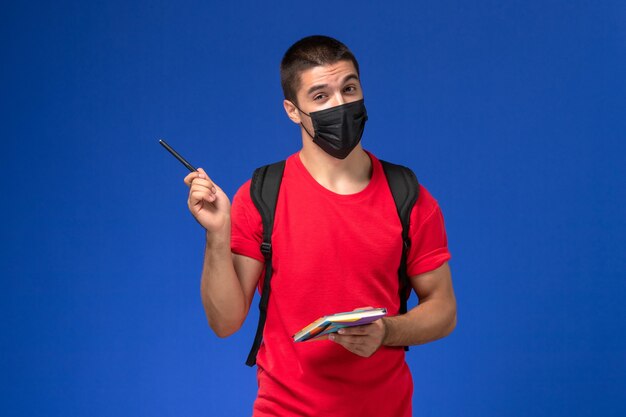 Vue de face étudiant masculin en t-shirt rouge portant un sac à dos en masque stérile noir tenant un stylo et un cahier sur le fond bleu.