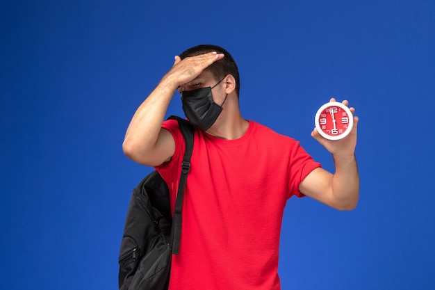 Vue de face étudiant masculin en t-shirt rouge portant un sac à dos avec masque tenant des horloges sur le fond bleu.