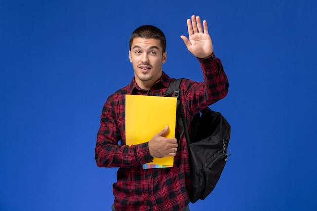 Vue de face de l'étudiant de sexe masculin en chemise à carreaux rouge avec sac à dos tenant des fichiers jaunes en agitant sur le mur bleu