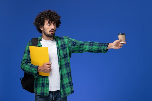 Vue de face de l'étudiant de sexe masculin en chemise à carreaux vert portant un sac à dos noir et tenant des fichiers et du café sur le mur bleu