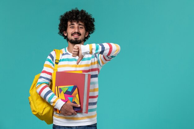 Vue de face de l'étudiant de sexe masculin portant un sac à dos jaune tenant des fichiers et des cahiers sur le mur bleu