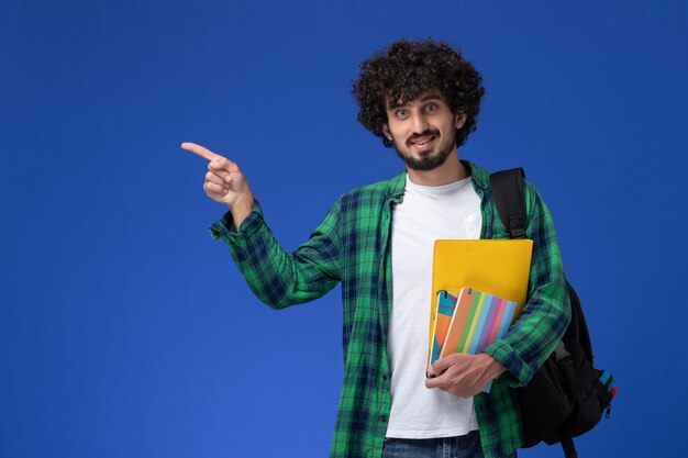 Vue de face de l'étudiant de sexe masculin portant un sac à dos noir tenant des cahiers et des fichiers sur le mur bleu