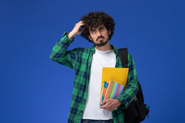 Vue de face de l'étudiant de sexe masculin portant un sac à dos noir tenant des cahiers et des fichiers pensant sur le mur bleu