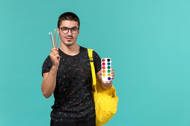 Vue de face de l'étudiant de sexe masculin en t-shirt foncé sac à dos jaune tenant des peintures et des glands sur le mur bleu clair
