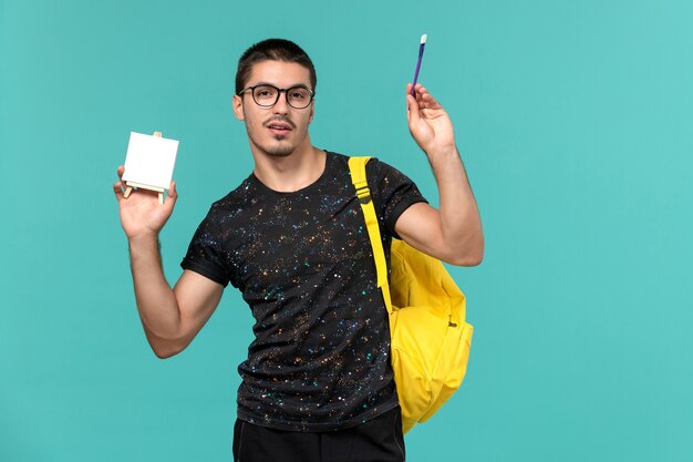 Vue de face de l'étudiant de sexe masculin en t-shirt foncé sac à dos jaune tenant pompon et chevalet sur le mur bleu clair