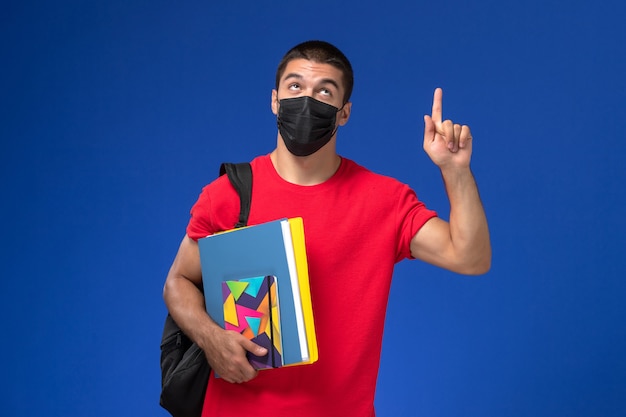 Vue de face étudiant de sexe masculin en t-shirt rouge portant sac à dos en masque stérile noir tenant des fichiers avec le doigt levé sur le fond bleu.