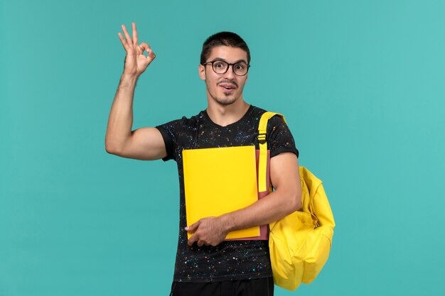 Vue de face de l'étudiant en t-shirt foncé sac à dos jaune tenant différents fichiers sur le mur bleu clair