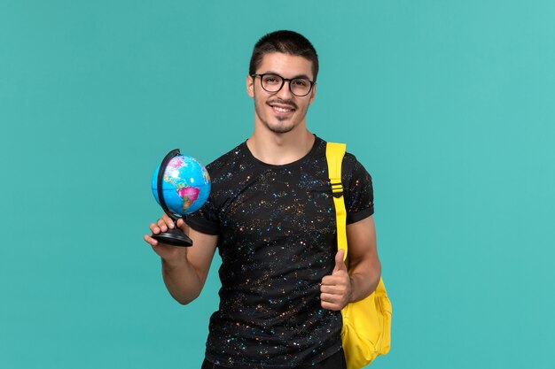 Vue de face de l'étudiant en t-shirt foncé sac à dos jaune tenant petit globe sur le mur bleu