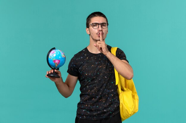 Vue de face de l'étudiant en t-shirt foncé sac à dos jaune tenant petit globe sur le mur bleu