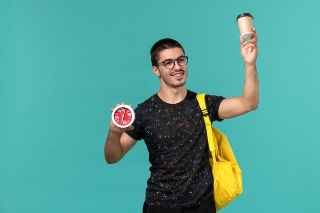 Vue de face de l'étudiant en t-shirt noir sac à dos jaune tenant du café et des horloges sur le mur bleu