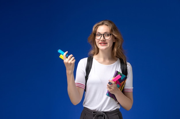 Vue de face de l'étudiante en chemise blanche portant un sac à dos et tenant un cahier avec des feutres sur le mur bleu