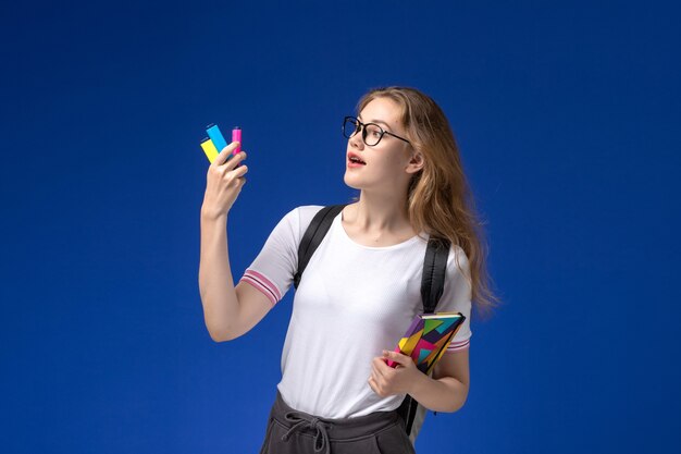 Vue de face d'une étudiante en chemise blanche portant un sac à dos et tenant un cahier sur le mur bleu