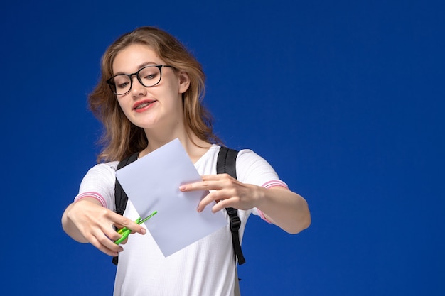 Vue de face de l'étudiante en chemise blanche portant un sac à dos et tenant du papier et des ciseaux sur le mur bleu