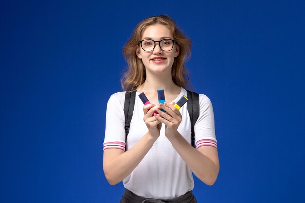 Vue de face d'une étudiante en chemise blanche portant un sac à dos et tenant des feutres sur le mur bleu