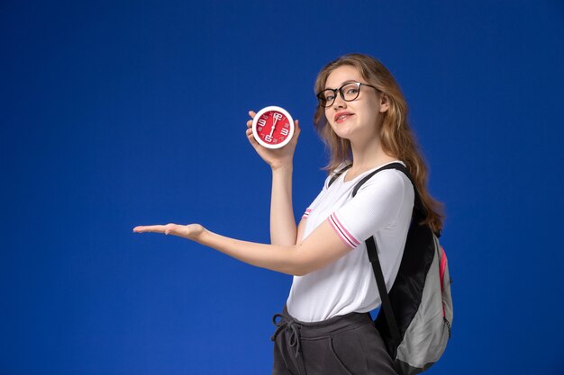 Vue de face de l'étudiante en chemise blanche portant un sac à dos et tenant des horloges sur le bureau bleu leçon d'université college female