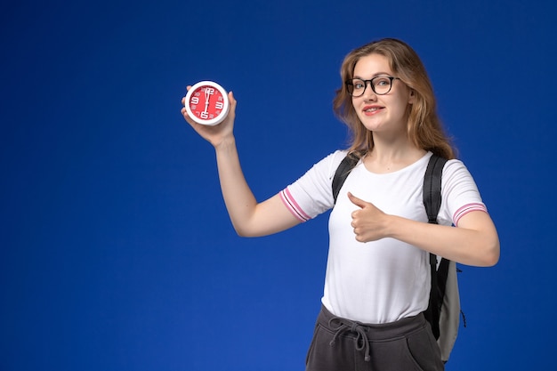 Vue de face de l'étudiante en chemise blanche portant sac à dos tenant des horloges sur le mur bleu