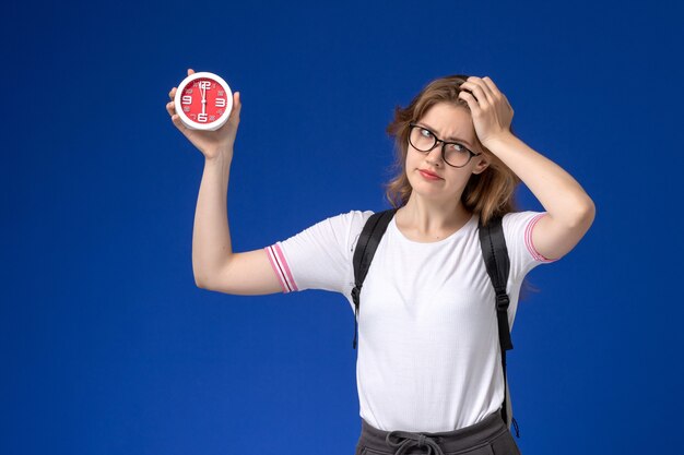 Vue de face de l'étudiante en chemise blanche portant sac à dos tenant des horloges sur le mur bleu