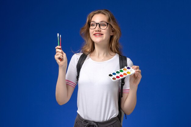 Vue de face d'une étudiante en chemise blanche portant un sac à dos et tenant de la peinture et des pinceaux