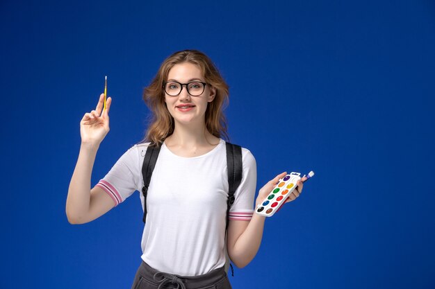 Vue de face de l'étudiante en chemise blanche portant un sac à dos et tenant un pinceau d'art souriant sur le mur bleu