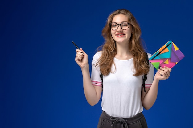 Vue de face de l'étudiante en chemise blanche tenant un stylo et un cahier sur le mur bleu