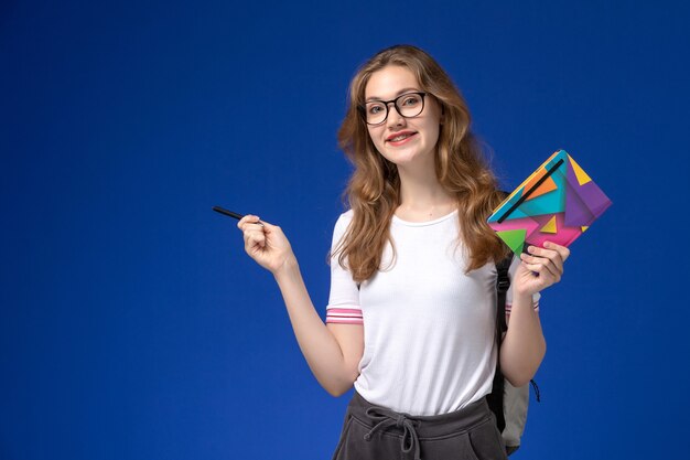 Vue de face de l'étudiante en chemise blanche tenant un stylo et un cahier sur le mur bleu