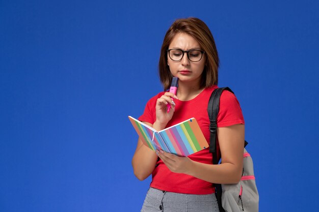 Vue de face de l'étudiante en chemise rouge avec sac à dos holding feutres lecture cahier sur le mur bleu
