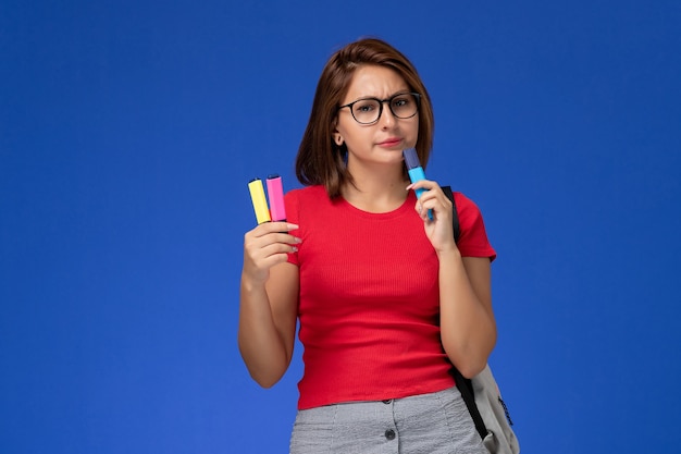 Vue de face de l'étudiante en chemise rouge avec sac à dos tenant des feutres pensant sur le mur bleu