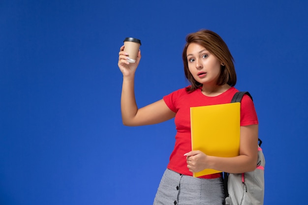 Vue de face de l'étudiante en chemise rouge avec sac à dos tenant des fichiers jaunes et café sur mur bleu