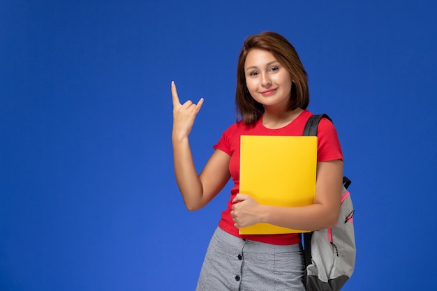 Vue de face de l'étudiante en chemise rouge avec sac à dos tenant des fichiers jaunes sur le mur bleu clair