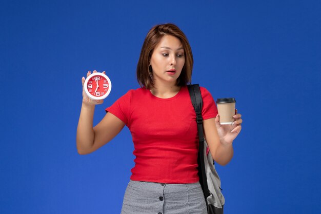 Vue de face de l'étudiante en chemise rouge avec sac à dos tenant des horloges et du café sur le mur bleu