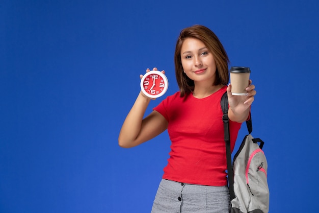 Vue de face de l'étudiante en chemise rouge avec sac à dos tenant des horloges et du café sur le mur bleu