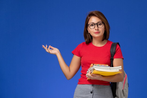 Vue de face de l'étudiante en chemise rouge avec sac à dos tenant des livres et des fichiers sur le mur bleu clair