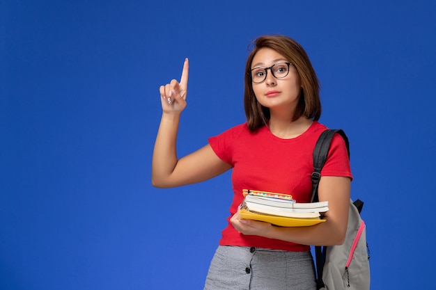 Vue de face de l'étudiante en chemise rouge avec sac à dos tenant des livres et des fichiers sur le mur bleu clair