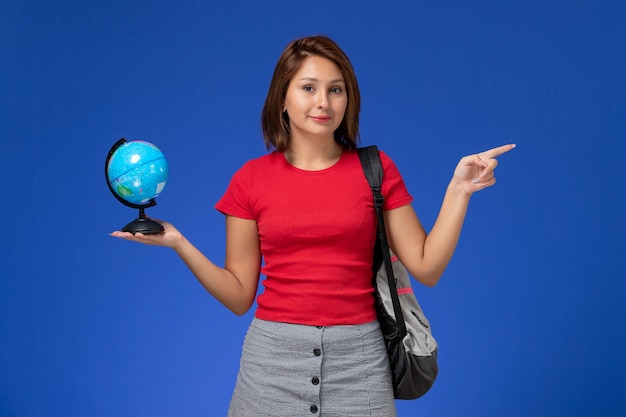 Vue de face de l'étudiante en chemise rouge avec sac à dos tenant petit globe sur mur bleu clair