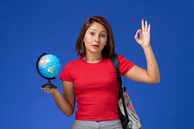 Vue de face de l'étudiante en chemise rouge avec sac à dos tenant petit globe sur le mur bleu