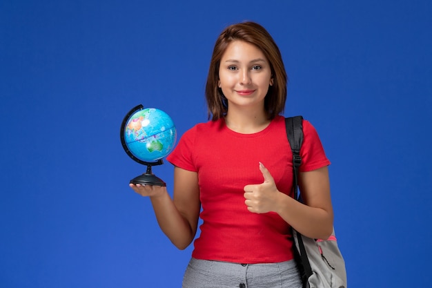 Vue de face de l'étudiante en chemise rouge avec sac à dos tenant petit globe souriant sur mur bleu