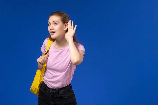 Vue de face de l'étudiante en t-shirt rose avec sac à dos jaune essayant d'entendre sur le mur bleu