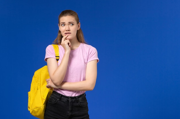 Vue de face de l'étudiante en t-shirt rose avec sac à dos jaune pensant sur le mur bleu