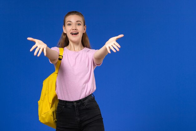 Vue de face de l'étudiante en t-shirt rose avec sac à dos jaune posant sur le mur bleu