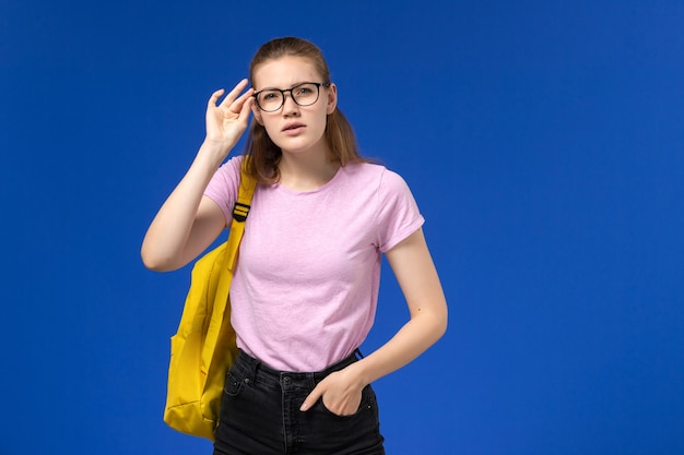 Vue de face de l'étudiante en t-shirt rose avec sac à dos jaune posant sur le mur bleu