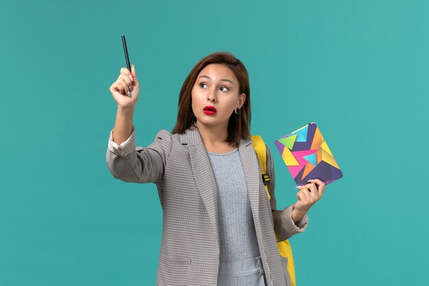 Vue de face de l'étudiante en veste grise portant un sac à dos jaune tenant un cahier et un stylo sur le mur bleu clair